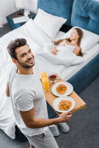 Над головой вид улыбающегося мужчины, держащего деревянный поднос с завтраком в то время как женщина спит в постели — стоковое фото