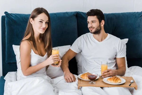 Young woman holding glass of orange juice while boyfriend sitting with wooden tray — Stock Photo