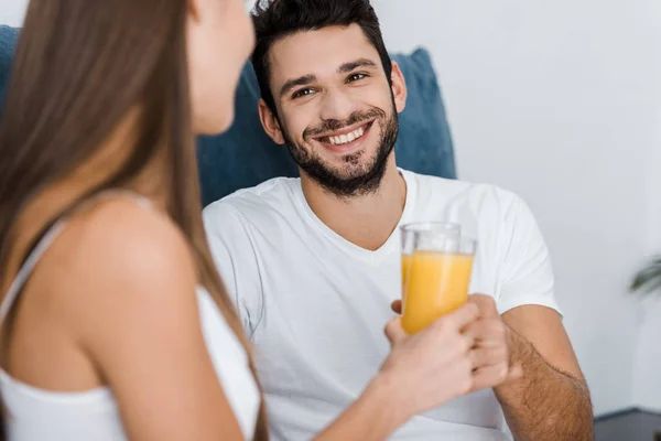 Foco seletivo de belo homem sorridente olhando para a namorada e segurando vidro de suco de laranja — Fotografia de Stock