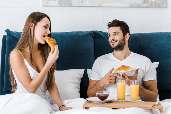Jeune couple manger des croissants savoureux pendant le petit déjeuner au lit — Photo de stock