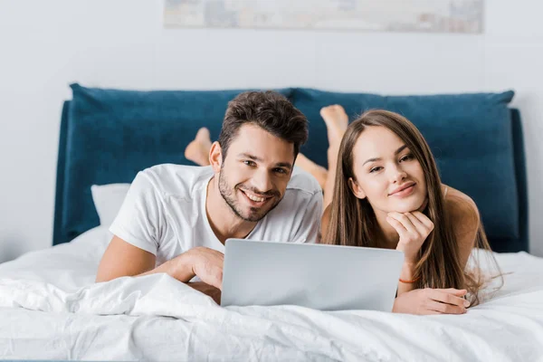 Jovem casal sorrindo deitado na cama com laptop e olhando para a câmera — Fotografia de Stock