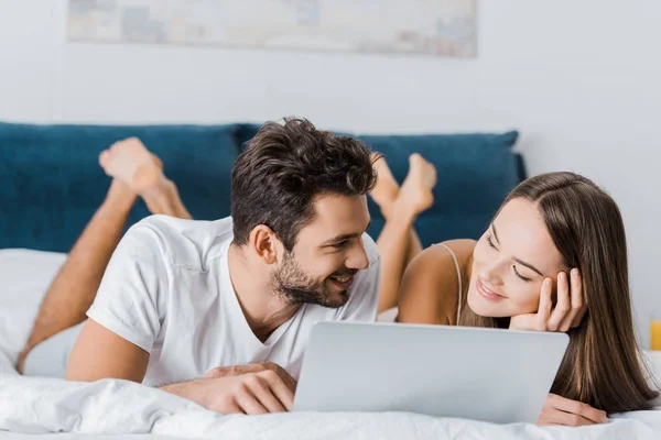 Joven sonriente pareja acostada en la cama y el uso de ordenador portátil - foto de stock