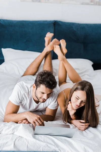 Jeune couple couché au lit avec les jambes croisées et à l'aide d'un ordinateur portable — Photo de stock