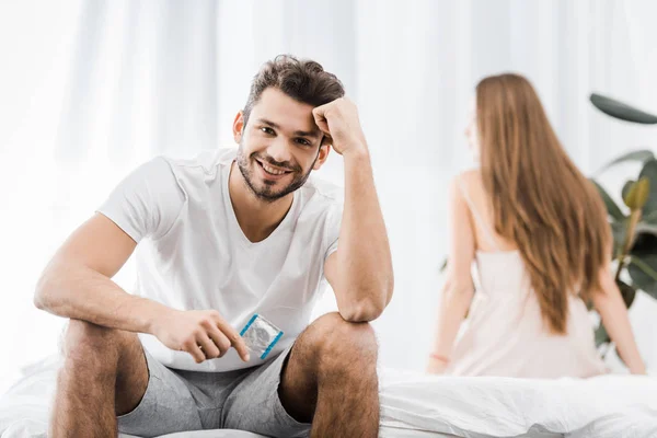 Cheerful handsome man sitting on bed and holding condom — Stock Photo