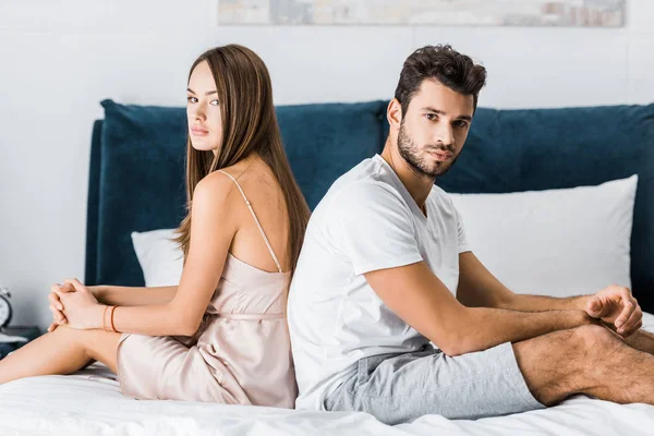 Young frustrated couple in pajamas sitting on bed back to back and looking at camera — Stock Photo