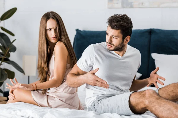 Несчастливая пара в пижаме сидит на кровати спиной к спине и жестикулирует сердито, сексуальные проблемы концепции — стоковое фото