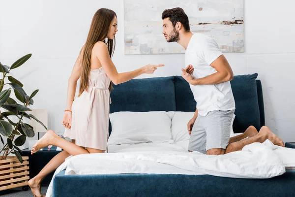 Young attractive woman poiting finger angrily on stressed gesturing man while standing on bed — Stock Photo