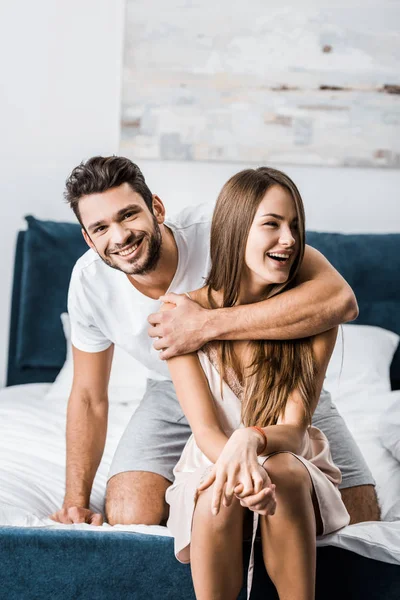 Joven hombre abrazando a la mujer con una mano y sonriendo mientras está sentado en la cama - foto de stock