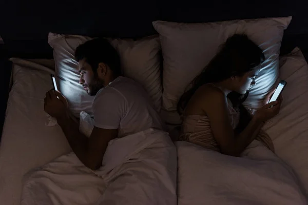 Vista superior de la joven pareja frustrada acostada en la cama por la noche y el uso de teléfonos inteligentes - foto de stock