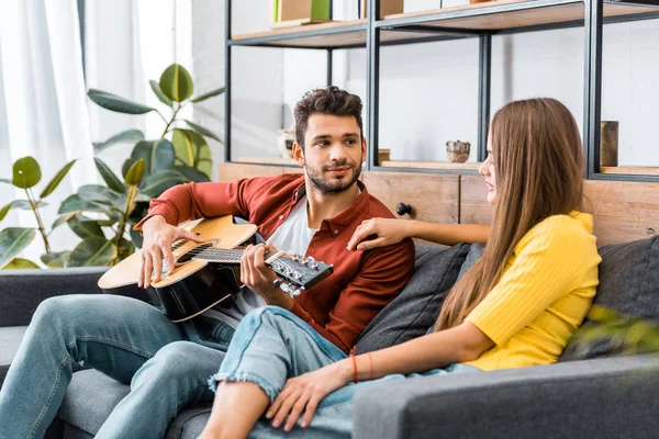 Joven hombre guapo sentado en el sofá en la sala de estar y tocando la guitarra acústica para la novia - foto de stock