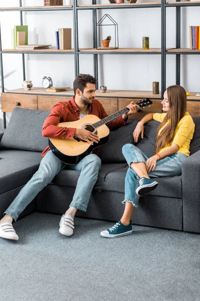 Молодий красивий чоловік грає на гітарі для дівчини у вітальні — стокове фото