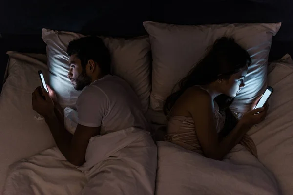Vista superior de la joven pareja acostada en la cama por la noche y utilizando teléfonos inteligentes - foto de stock