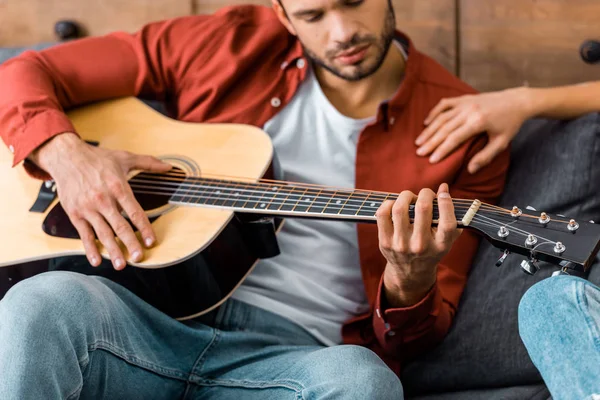 Cropped view of young handsome man playing acoustic guitar while sitting on sofa — Stock Photo