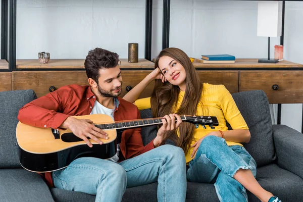 Junger schöner Mann sitzt auf Sofa im Wohnzimmer und spielt Gitarre für attraktive Freundin — Stockfoto
