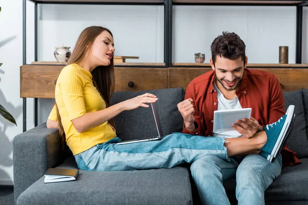 Молодая пара, сидящая на диване в гостиной, проводит время с ноутбуком и цифровой планшет — стоковое фото
