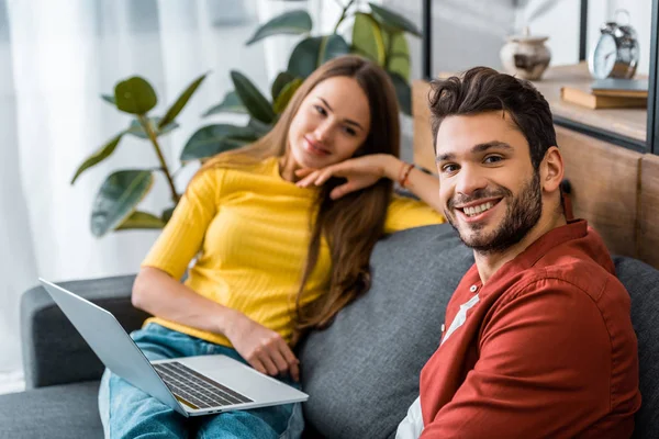 Joven feliz pareja sentado en el sofá con el ordenador portátil y sonriendo mientras mira a la cámara - foto de stock