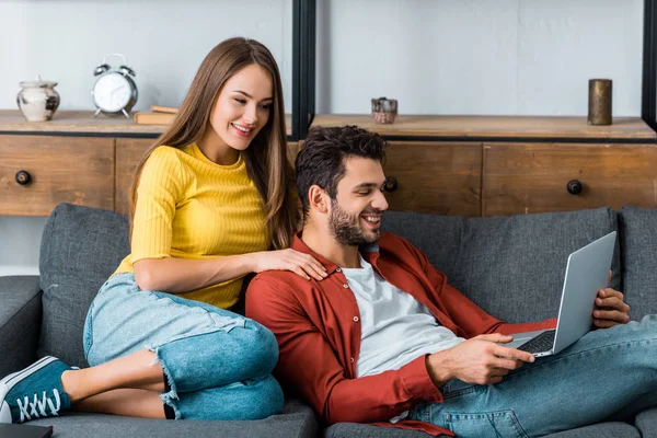 Jovem casal feliz sentado no sofá com laptop e sorrindo enquanto olha para a tela — Fotografia de Stock