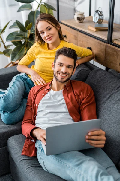 Junges attraktives Paar sitzt mit Laptop auf dem Sofa und lächelt in die Kamera — Stockfoto