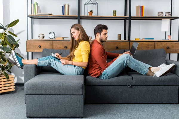 Giovane donna sorridente con taccuino seduta schiena a schiena sul divano con uomo utilizzando laptop — Foto stock