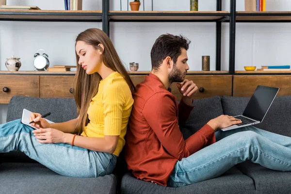 Young couple sitting back to back on sofa using laptop and writing in notebook — Stock Photo