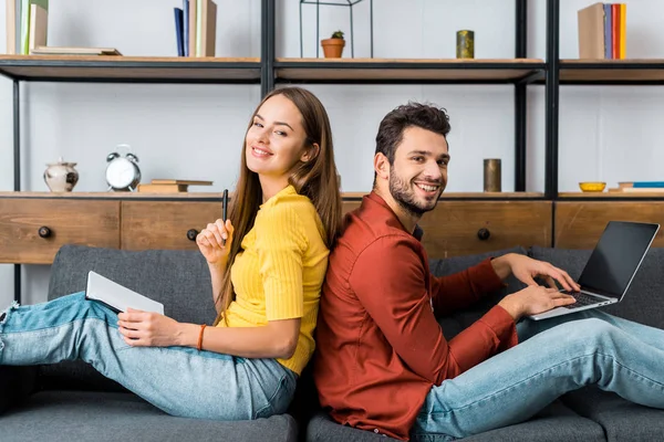 Junges glückliches Paar sitzt mit Notizbuch und Laptop im Wohnzimmer und lächelt in die Kamera — Stockfoto