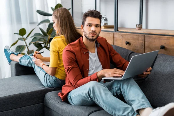 Young man holding laptop and sitting back to back with woman on sofa — Stock Photo
