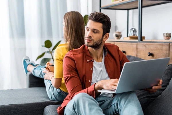 Schöner bärtiger Mann sitzt auf Sofa und benutzt Laptop in der Nähe Freundin — Stockfoto