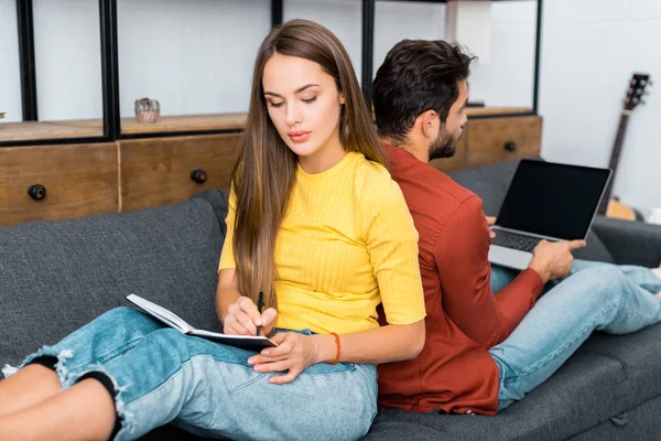 Mujer pensativa escribiendo en cuaderno y sentado cerca de novio con portátil - foto de stock