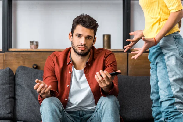 Frustrado hombre sentado en el sofá y mostrando gesto encogiéndose de hombros cerca de novia enojada - foto de stock