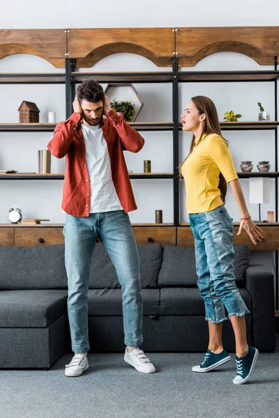 Angry girlfriend screaming at boyfriend while man putting hands on ears in living room — Stock Photo