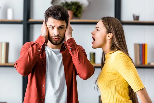 Selective focus of angry girlfriend screaming at boyfriend while man putting hands on ears — Stock Photo