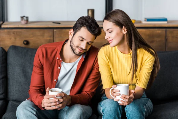 Happy couple sitting on sofa and holding cups with drinks — Stock Photo