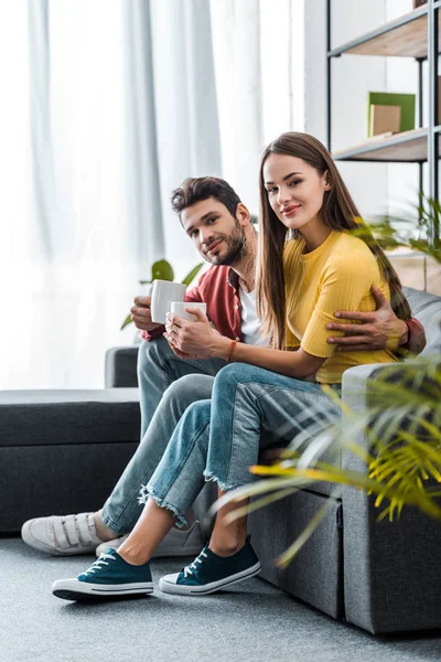 Glückliches Paar sitzt auf Sofa mit Tassen — Stockfoto