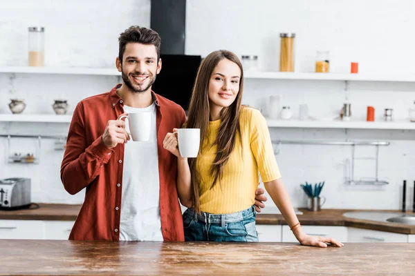 Coppia allegra in piedi in cucina e sorridente con tazze — Foto stock