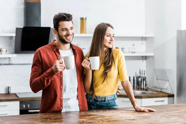Cheerful couple standing in kitchen with cups — Stock Photo