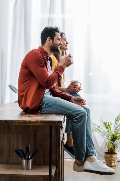 Vista lateral de la pareja sentada en una mesa de madera en la cocina y sosteniendo tazas - foto de stock