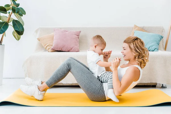 Mujer sosteniendo hijo pequeño y entrenamiento en la estera de fitness en la sala de estar - foto de stock