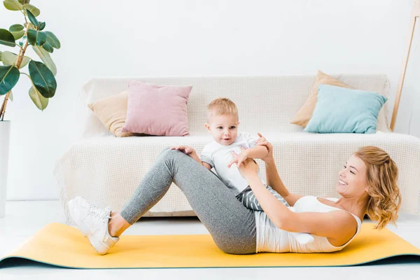 Frau hält Kleinkindsohn und trainiert zu Hause auf Fitnessmatte — Stockfoto