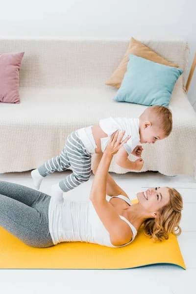 Mother lying on fitness mat and holding toddler child over head — Stock Photo