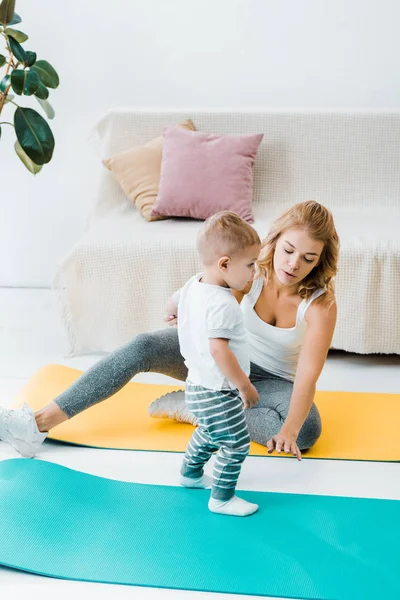 Madre sentada en la colchoneta de fitness y hablando con el lindo hijo bebé - foto de stock