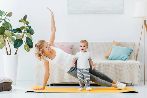 Mulher deitada no tapete de fitness e sorrindo para menino adorável — Fotografia de Stock