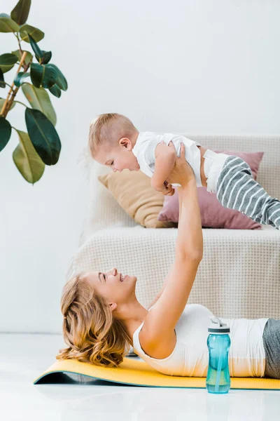 Madre sdraiata sul tappeto fitness e giocare con adorabile figlio vicino bottiglia d'acqua — Foto stock