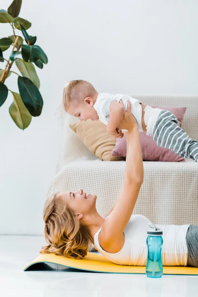 Mãe deitada no tapete de fitness e brincando com o filho adorável perto de garrafa de água — Fotografia de Stock