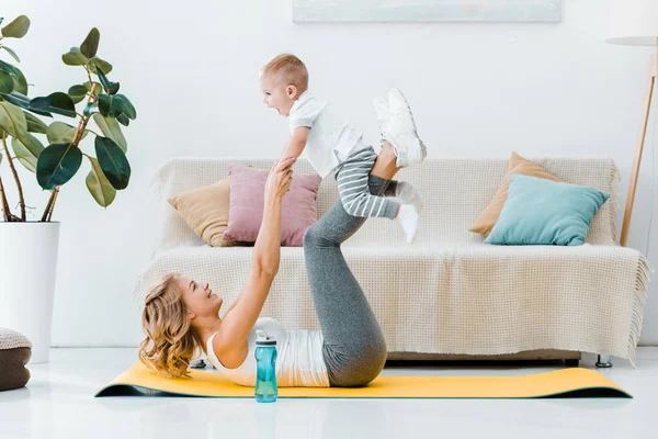 Femme couchée sur tapis de fitness, levant les yeux et élevant adorable enfant avec les pieds — Photo de stock