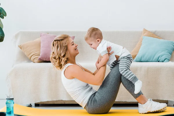 Mujer acostada en la alfombra de fitness mientras entrena y sostiene a un niño adorable en las manos - foto de stock