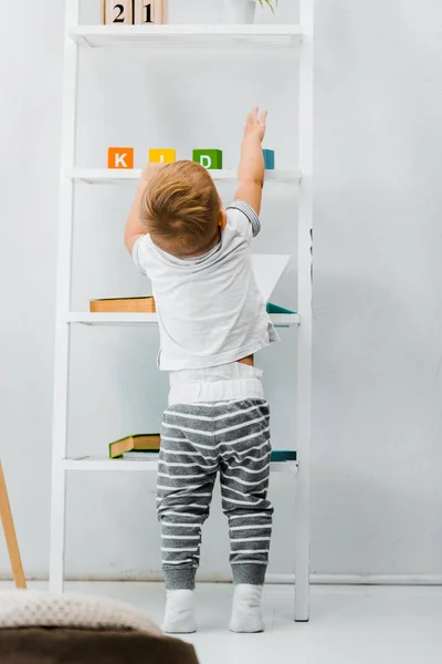 Cute toddler boy standing near rack and reaching for toys on shelves — Stock Photo
