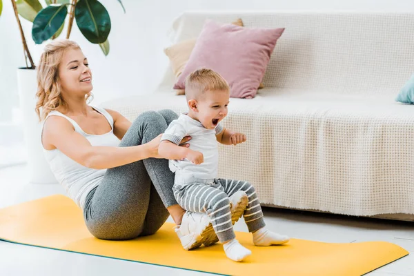 Madre seduta sul tappeto fitness mentre solleva il bambino in aria — Foto stock