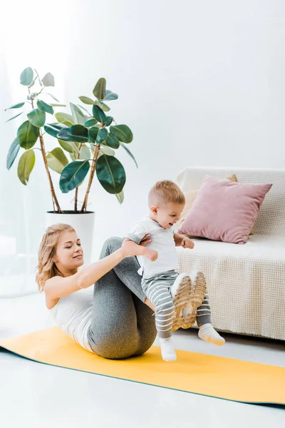 Madre haciendo ejercicio en la alfombra de fitness y jugando con su hijo - foto de stock