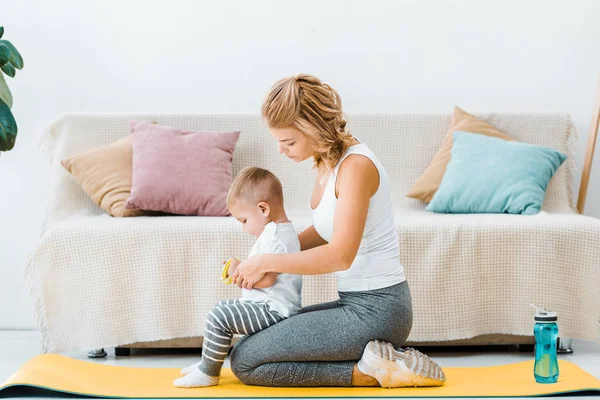 Donna seduta sul tappetino fitness e con in mano un bambino in salotto — Foto stock