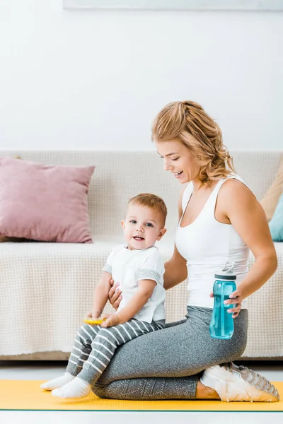 Smiling cute toddler boy and attractive woman sitting on fitness mat and holding sports bottle — Stock Photo